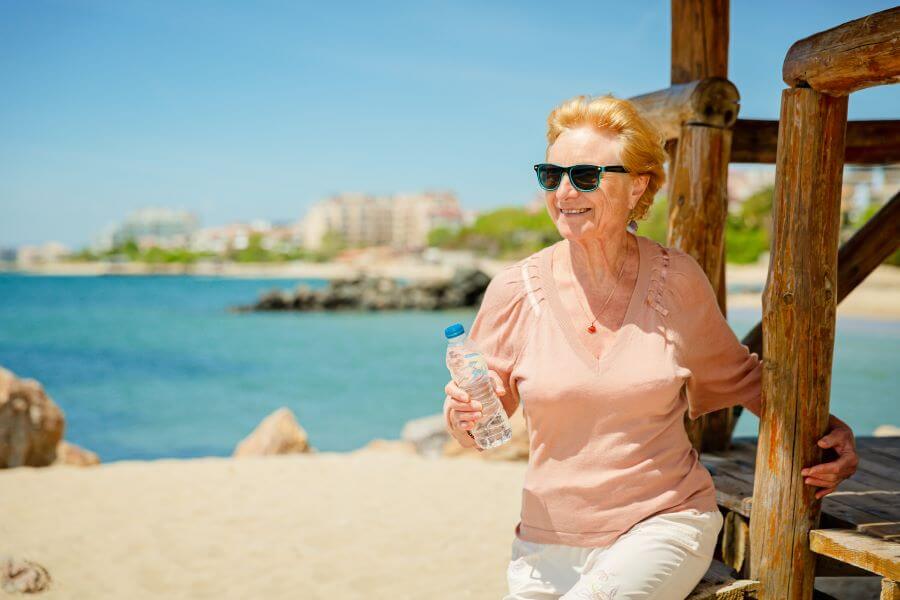 an elderly woman sits on a beach holding a bottle of water